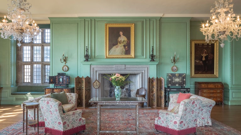 Drawing room with floral chairs and table set around a large fire place. The walls are green and there are two chandeliers hanging from the ceiling